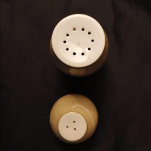 May include: Overhead shot of two ceramic salt and pepper shakers. The shakers are beige with white tops. The top shaker has 12 holes, while the bottom shaker has 3 holes. The background is a solid black.
