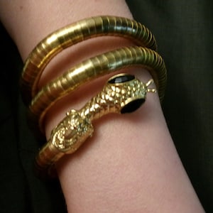 May include: A gold snake bracelet with a black gem on the head. The bracelet is made of multiple coiled bands.