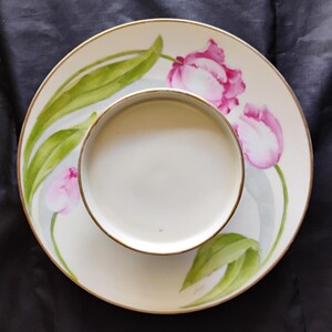 May include: A white ceramic plate and bowl set with a gold rim. The plate features a hand-painted design of pink tulips and green leaves. The bowl sits centered on the plate, creating a cohesive dining set.