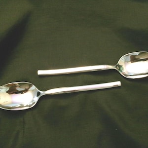 May include: Two silver spoons with a shiny finish. The spoons are laid out on a dark green surface.