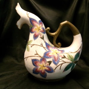 May include: A white ceramic teapot shaped like a bird with a gold handle. The teapot is decorated with blue, purple, and yellow flowers.