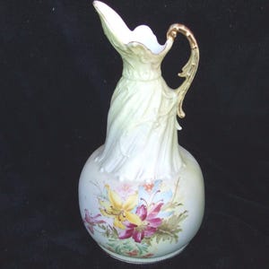 May include: A vintage porcelain pitcher with a bulbous base and a slender, fluted neck. The pitcher is primarily cream-colored with a gold-colored handle and floral designs in yellow, pink, and blue. The pitcher measures approximately 10 inches in height.