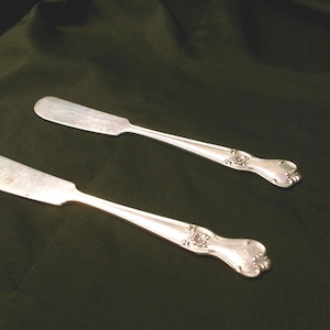 May include: Two silver butter knives with ornate handles. The knives have a slightly tarnished finish.