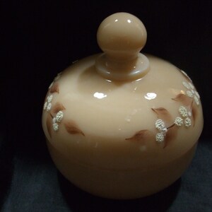 May include: A beige glass jar with a lid and a floral design. The jar has a white floral pattern with brown leaves.