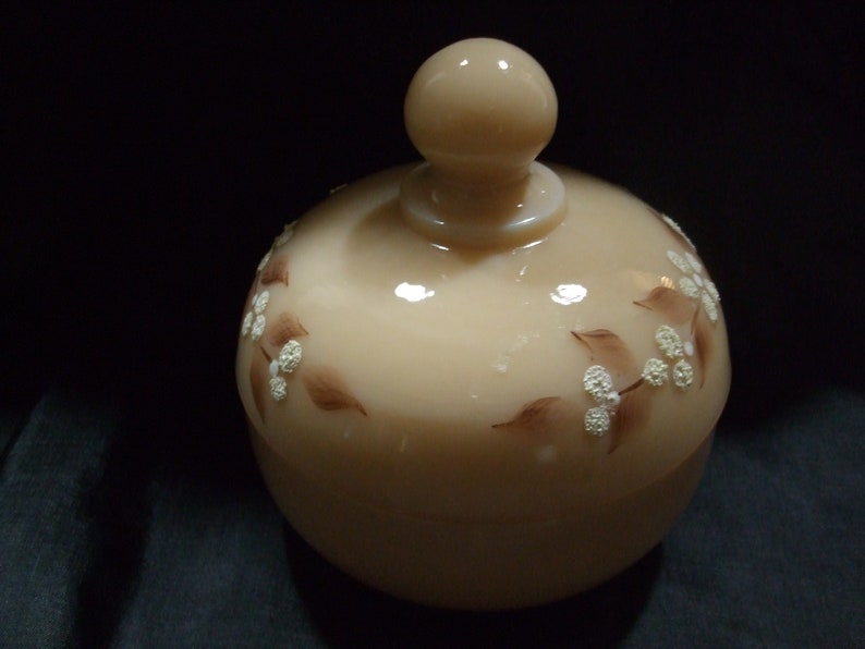 May include: A beige glass jar with a lid and a floral design. The jar has a white floral pattern with brown leaves.