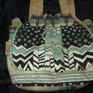 May include: A beige canvas tote bag with a design of three black, white, and green cats with blue eyes. The cats are stylized and have a geometric pattern. The bag has a brown leather handle.