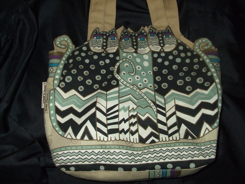 May include: A beige canvas tote bag with a design of three black, white, and green cats with blue eyes. The cats are stylized and have a geometric pattern. The bag has a brown leather handle.