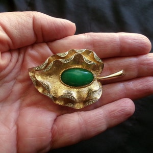 May include: A gold-tone brooch in the shape of a leaf with a green cabochon stone in the center. The leaf is decorated with small rhinestones around the edge.