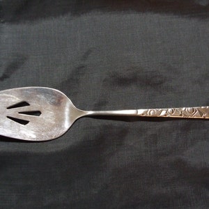 May include: A silver-toned metal cake server featuring a wide, pointed blade with three triangular cutouts and a flat handle decorated with a repeating geometric pattern of embossed squares and circles along its entire length.