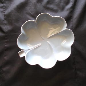 May include: A white, clover-shaped serving dish. The dish has four rounded lobes, each with a heart-like shape. The dish is made of a glossy material and sits on a dark background. The dish is a decorative item.