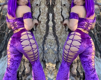 May include: Purple velvet bell bottom pants with cutouts and lace-up details. The pants are worn by a person with purple hair and are styled with a matching purple crop top.