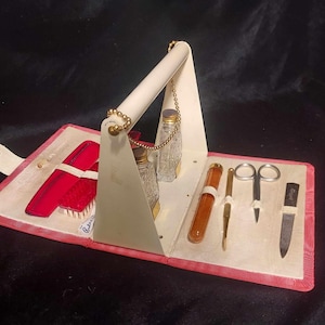 May include: A vintage pink and white travel vanity case with a handle, containing a comb, brush, scissors, tweezers, a nail file, and two small glass bottles.
