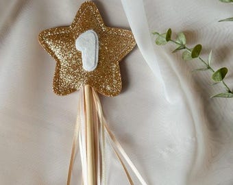 May include: A gold glitter star-shaped wand with the number 1 in white. The wand has a wooden stick handle and is adorned with flowing ribbons in cream and gold. This festive item is likely for a birthday celebration.