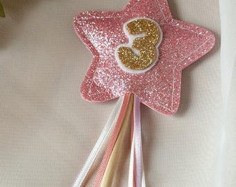 May include: A pink glitter star-shaped wand with a gold glitter number 3 in the center. The wand has a wooden handle and colorful ribbons in white, pink, and lavender. A festive party decoration.