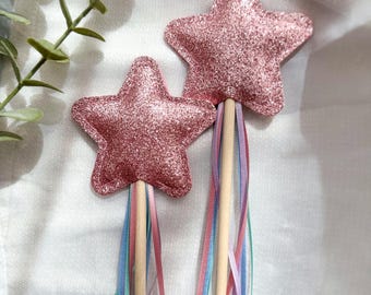 Puede incluir: Dos varitas mágicas de estrella rosa brillante con mangos de madera y cintas de colores. Las partes superiores en forma de estrella están cubiertas de purpurina rosa. Las varitas tienen cintas pastel en rosa, azul y lavanda.