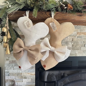 May include: Two fish-shaped Christmas stockings, one beige and one off-white, each with a burlap bow and a small tag with a name. They hang in front of a fireplace, decorated with pine branches and bells, creating a festive scene.