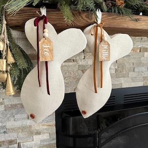 May include: Two beige fish-shaped Christmas stockings with wooden tags that say "Leo" and "Millie". The stockings are hung on a wooden mantel with a garland of pine branches.