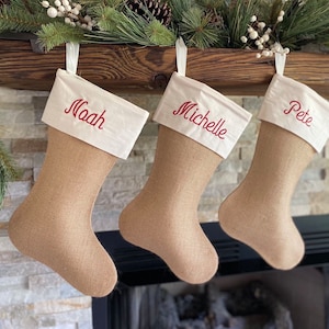 Puede incluir: Tres calcetines de Navidad de arpillera con puños de fieltro blanco y nombres bordados en rojo: Noah, Michelle y Pete.
