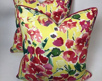 May include: Two decorative throw pillows with a vibrant floral pattern. The pillows feature a bright yellow background with red, pink, and green watercolor-style flowers. A dark red trim outlines the edges of the pillows, adding a contrasting detail.