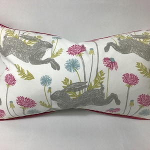 May include: A white lumbar pillow with a red border features a repeating pattern of gray hares leaping through a field of pink, blue, and green wildflowers.