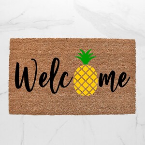 May include: A brown coir doormat with the word "Welcome" in black cursive lettering and a yellow pineapple with green leaves in the center.