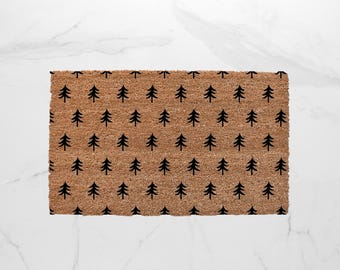 May include: A rectangular doormat with a natural brown background and a repeating pattern of black pine trees. The mat is made of a coarse, textured material.