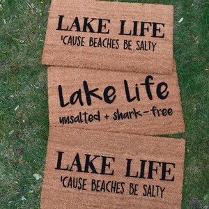 Puede incluir: Tres felpudos de fibra de coco marr&oacute;n con el texto "LAKE LIFE 'CAUSE BEACHES BE SALTY" en los felpudos superior e inferior y "Lake Life unsalted + shark-free" en el felpudo del medio.