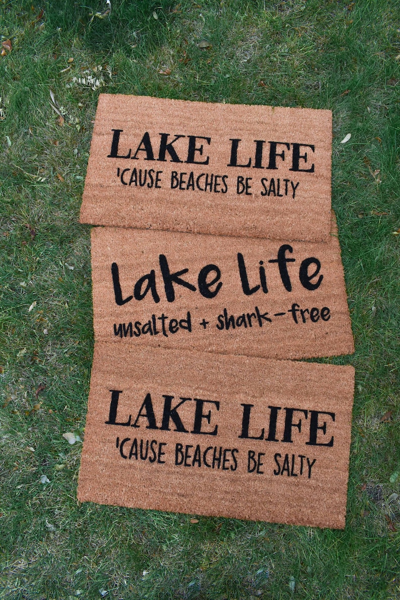 Puede incluir: Tres felpudos de fibra de coco marr&oacute;n con el texto "LAKE LIFE 'CAUSE BEACHES BE SALTY" en los felpudos superior e inferior y "Lake Life unsalted + shark-free" en el felpudo del medio.