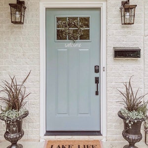Puede incluir: Una puerta delantera azul claro con una corona en la ventana y un letrero de bienvenida. La puerta tiene una manija de puerta negra y un buz&oacute;n negro en la pared a la derecha de la puerta. Una alfombra de bienvenida con el texto "Lake Life 'Cause Beaches Be Salty" est&aacute; en el suelo frente a la puerta. Dos urnas negras con plantas est&aacute;n a ambos lados de la puerta.