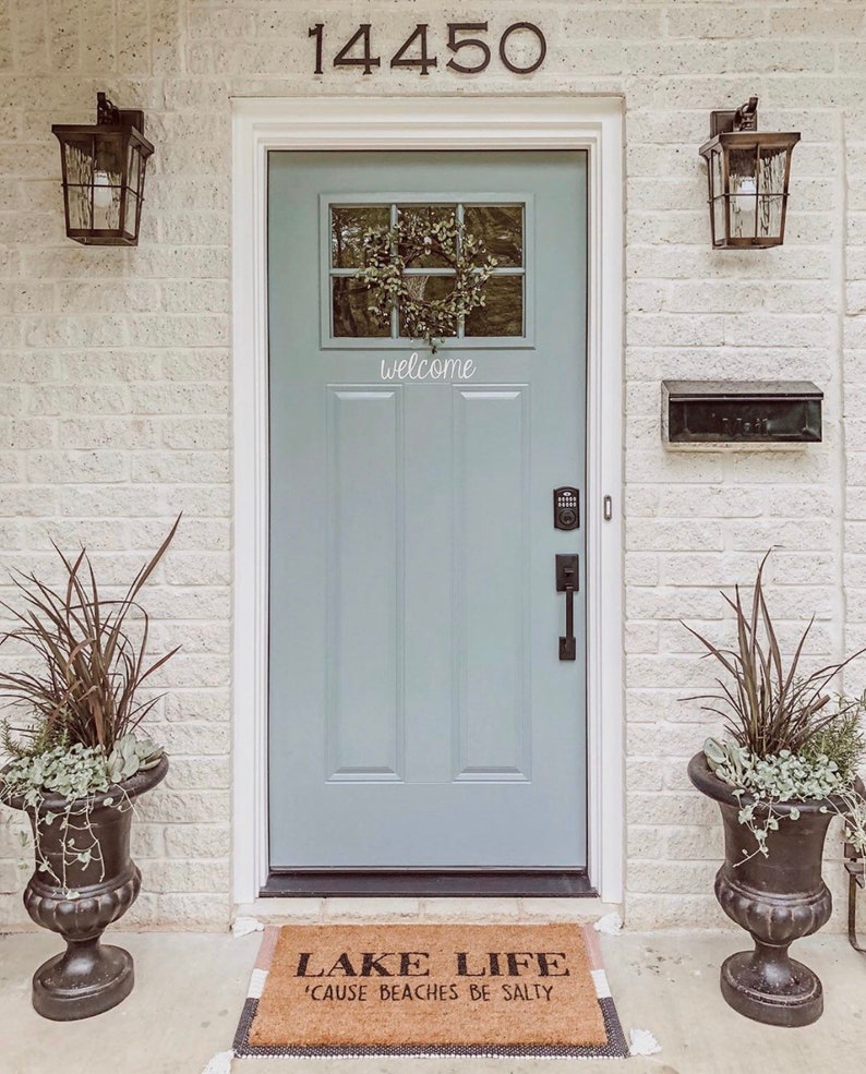 Puede incluir: Una puerta delantera azul claro con una corona en la ventana y un letrero de bienvenida. La puerta tiene una manija de puerta negra y un buz&oacute;n negro en la pared a la derecha de la puerta. Una alfombra de bienvenida con el texto "Lake Life 'Cause Beaches Be Salty" est&aacute; en el suelo frente a la puerta. Dos urnas negras con plantas est&aacute;n a ambos lados de la puerta.