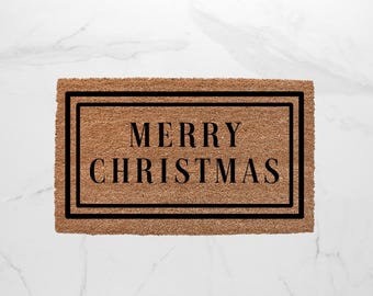 May include: A rectangular doormat made of natural coir fiber. The mat features the words "MERRY CHRISTMAS" in a bold, black font, framed by a black border. The background is a light brown color.