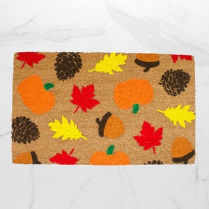 May include: A rectangular coir doormat featuring a repeating pattern of autumn-themed elements including orange pumpkins with green stems, bright red maple leaves, yellow oak leaves, brown pinecones, and brown acorns with dark brown caps.