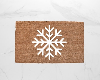 May include: A rectangular doormat made of brown coir fiber. A large white snowflake design is centered on the mat. The mat is placed on a white marble surface.