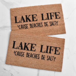 Puede incluir: Dos felpudos marrones con el texto "LAKE LIFE 'CAUSE BEACHES BE SALTY" en letras negras.