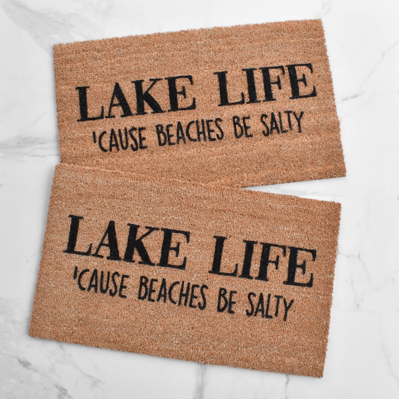 Puede incluir: Dos felpudos marrones con el texto "LAKE LIFE 'CAUSE BEACHES BE SALTY" en letras negras.