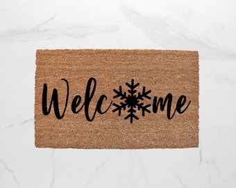 May include: A rectangular doormat made of natural coir fiber. The mat features the word "Welcome" in a cursive font, with a black snowflake replacing the "o" in "Welcome" and the "o" in "me".