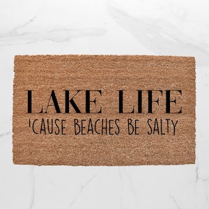 Puede incluir: Una alfombra de coco marr&oacute;n con el texto "LAKE LIFE 'CAUSE BEACHES BE SALTY" en negro.