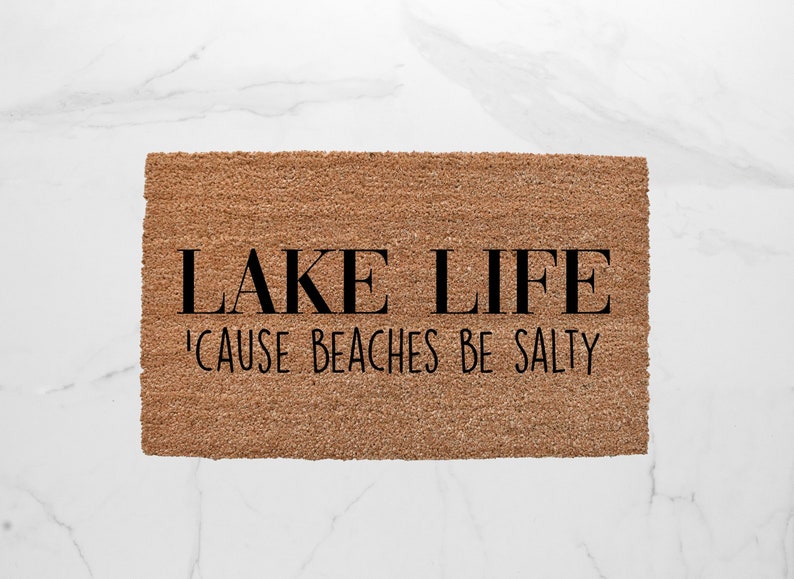 Puede incluir: Una alfombra de coco marr&oacute;n con el texto "LAKE LIFE 'CAUSE BEACHES BE SALTY" en negro.