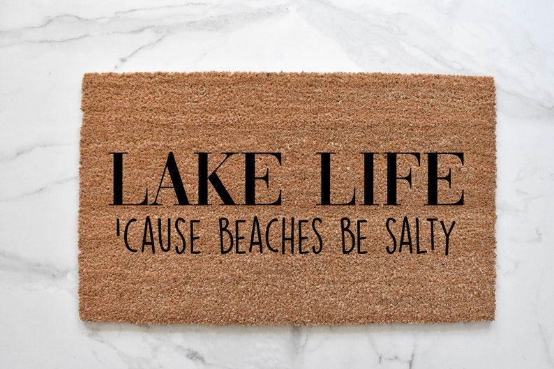 Puede incluir: Una alfombra de coco marr&oacute;n con el texto "LAKE LIFE 'CAUSE BEACHES BE SALTY" en negro.