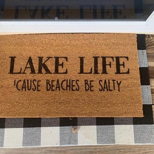 Puede incluir: Una alfombra de coco marr&oacute;n con el texto "LAKE LIFE 'CAUSE BEACHES BE SALTY" en letras negras. La alfombra est&aacute; sobre una alfombra a cuadros blancos y negros.