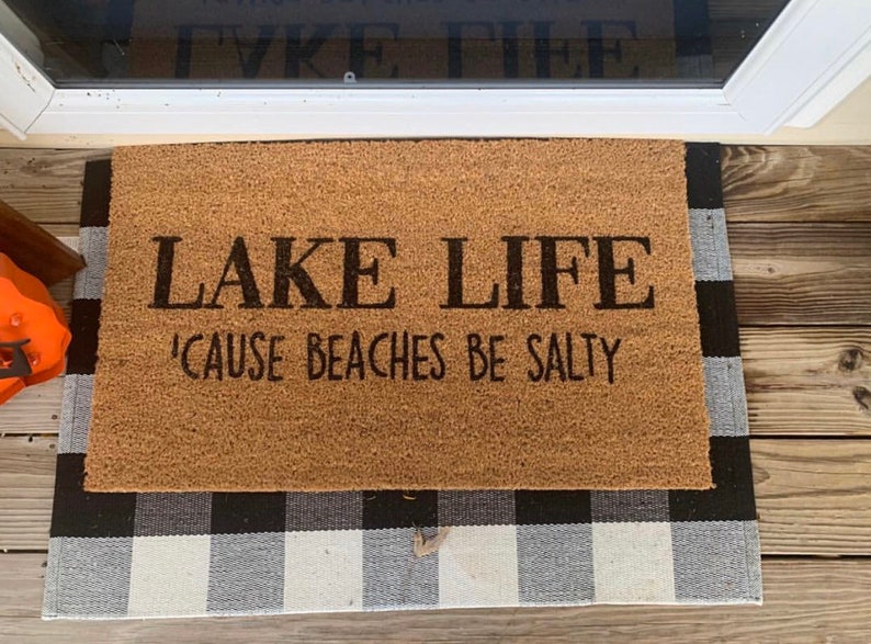 Puede incluir: Una alfombra de coco marr&oacute;n con el texto "LAKE LIFE 'CAUSE BEACHES BE SALTY" en letras negras. La alfombra est&aacute; sobre una alfombra a cuadros blancos y negros.