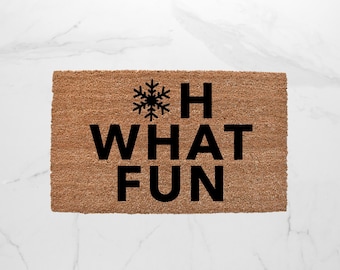 May include: A rectangular coir doormat with the phrase "OH WHAT FUN" in large, black capital letters. A black snowflake graphic is above the "H". The mat is a natural brown color and sits on a white marble surface.