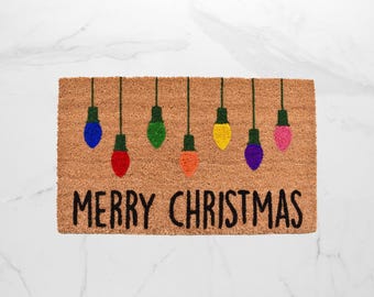 May include: A rectangular doormat made of natural coir fiber. It features a string of colorful Christmas lights in blue, green, red, orange, yellow, purple, and pink. The words "MERRY CHRISTMAS" are written in black.