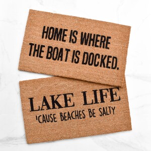 Puede incluir: Dos felpudos de fibra de coco marr&oacute;n con texto negro. El felpudo superior dice "HOME IS WHERE THE BOAT IS DOCKED." El felpudo inferior dice "LAKE LIFE 'CAUSE BEACHES BE SALTY."