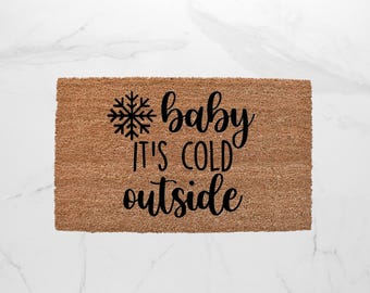 May include: A rectangular doormat made of natural brown coir fiber. The mat features a black snowflake graphic and the phrase "baby it's cold outside" in a stylish, cursive font, perfect for winter decor.