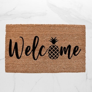 May include: A rectangular doormat made of natural brown coir fiber. The word "Welcome" is written in a black script font, with a black pineapple graphic replacing the "o" in "Welcome".