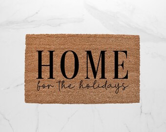 May include: Rectangular doormat made of natural brown coir fiber. The mat features the word "HOME" in large, bold, black capital letters, with "for the holidays" in a smaller, cursive font below. The mat is placed on a white marble surface.