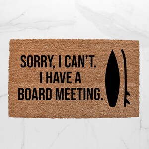 May include: Rectangular doormat made of natural coir fiber. The mat features the text "SORRY, I CAN'T. I HAVE A BOARD MEETING." in black, along with a black surfboard silhouette. The background is a white marble.