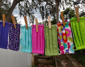 May include: Eight fabric face masks hanging on a clothesline with wooden clothespins. The masks are in various colors and patterns, including purple, teal, pink, green, and floral.