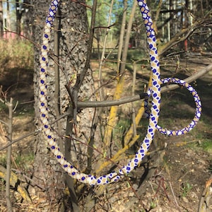 Puede incluir: Un conjunto de collar y pulsera de cuentas azules, blancas y doradas. El collar est&aacute; drapeado sobre una rama y la pulsera est&aacute; en el suelo.